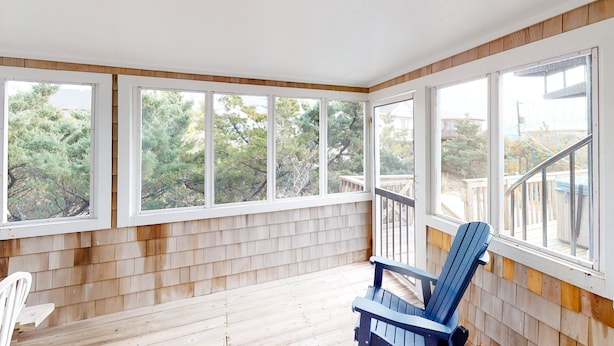 A covered porch with wooden flooring, a blue Adirondack chair, and large windows offering a view of trees and a deck. - Twin Tides #14-AC: Semi-Oceanfront, Dog-Friendly, Shared Pool, Hot Tub (Avon)