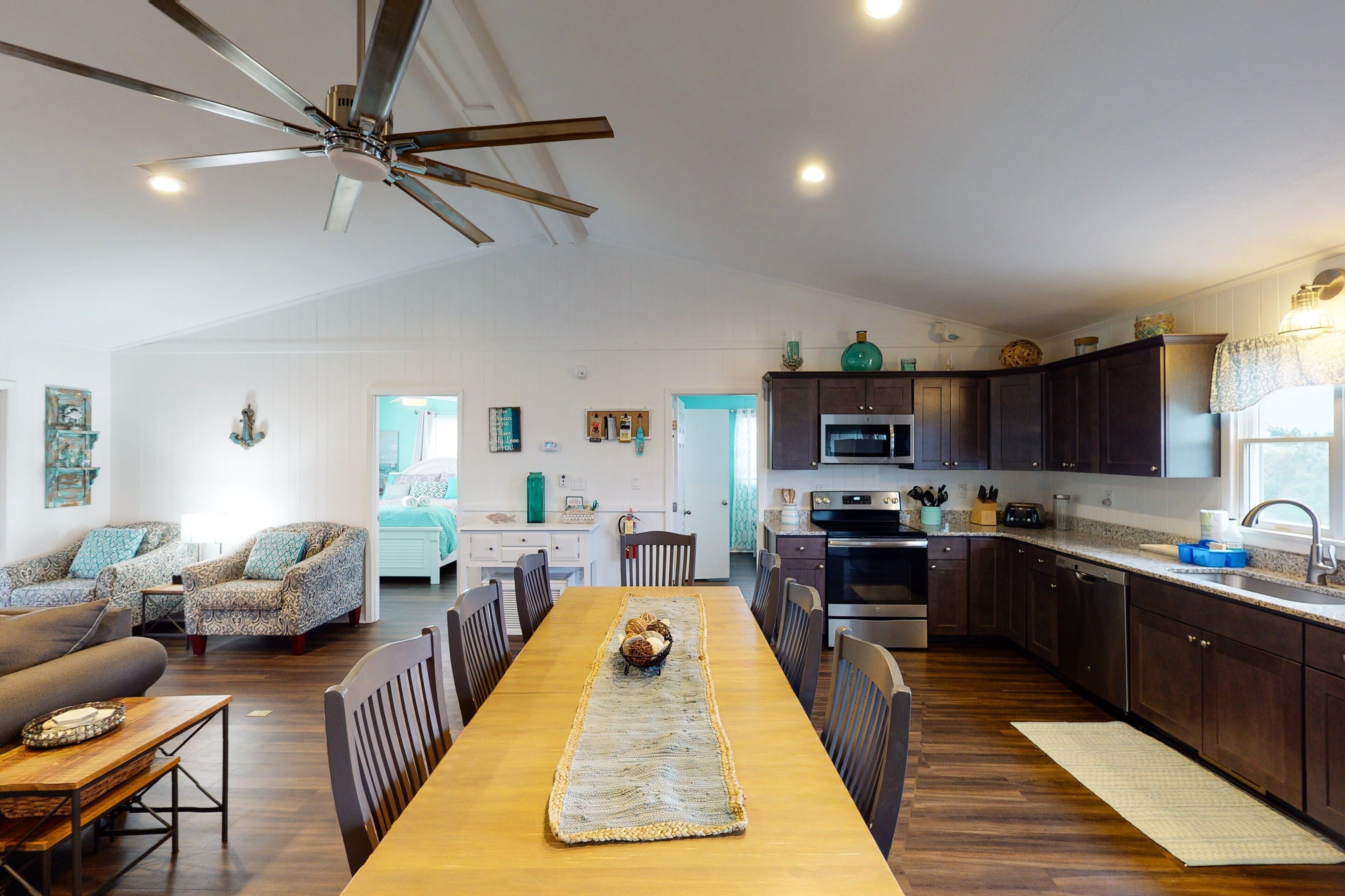 A spacious living room with a long wooden dining table, a ceiling fan, and a kitchen area with dark cabinets and stainless steel appliances.