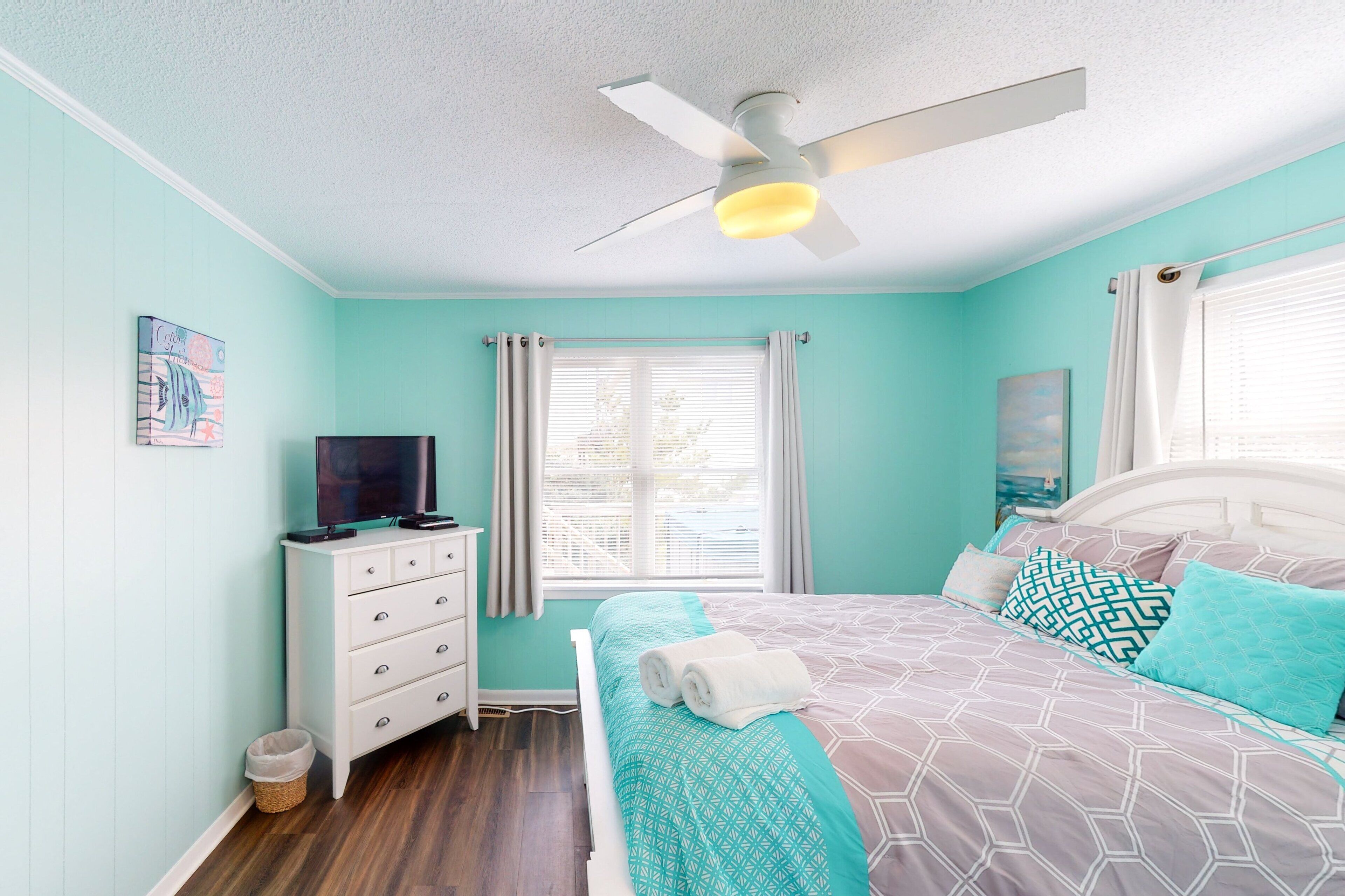 A bedroom with a bed, a ceiling fan, a television on a dresser, and a window with curtains.
