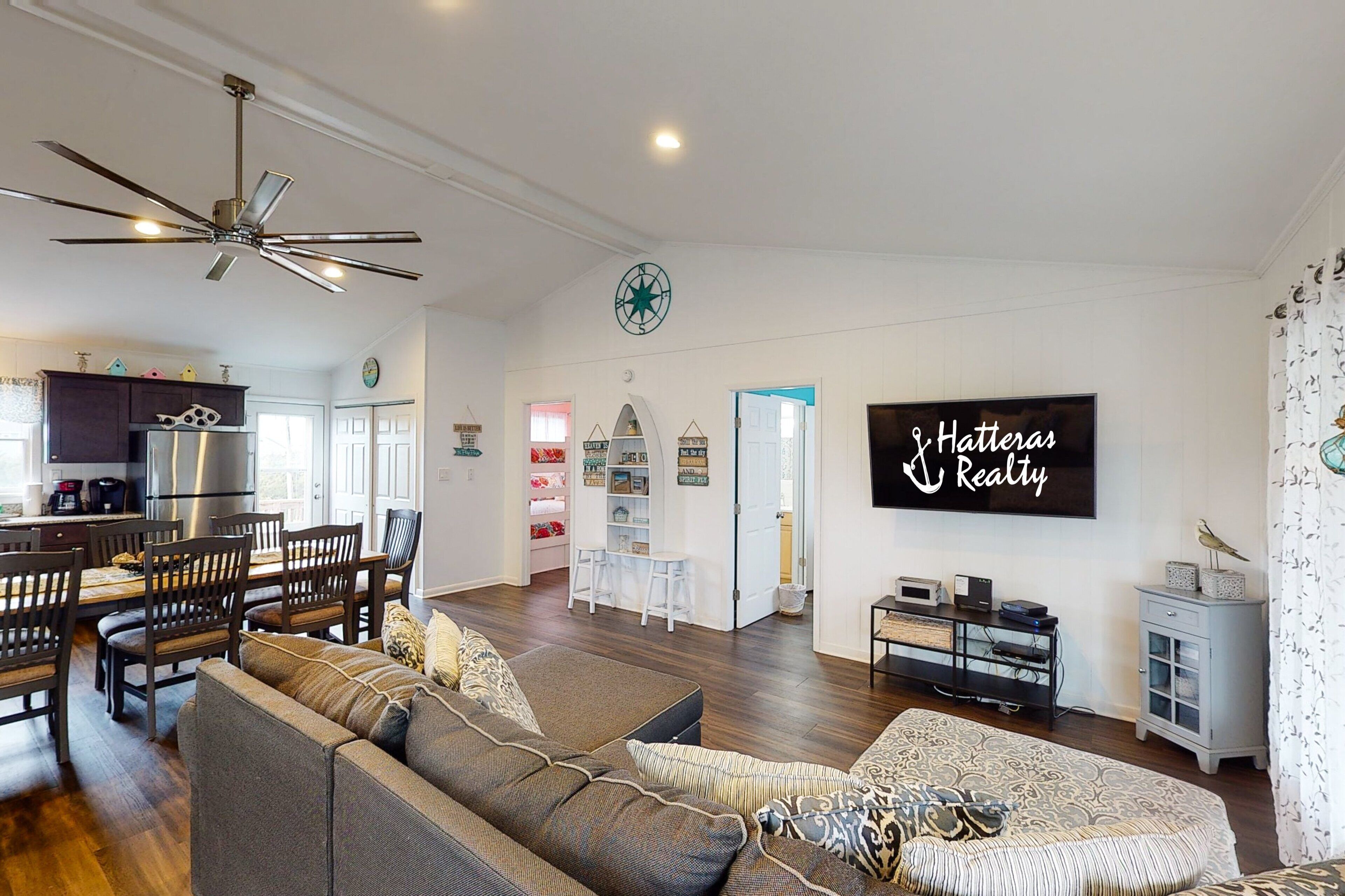 A spacious living room with a large sofa, a dining area with wooden chairs, a kitchen with modern appliances, and a ceiling fan.
