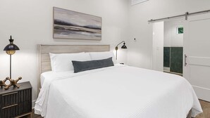 A neatly made bed with a white comforter and a dark pillow, a bedside lamp, a small wooden nightstand, and a painting on the wall.