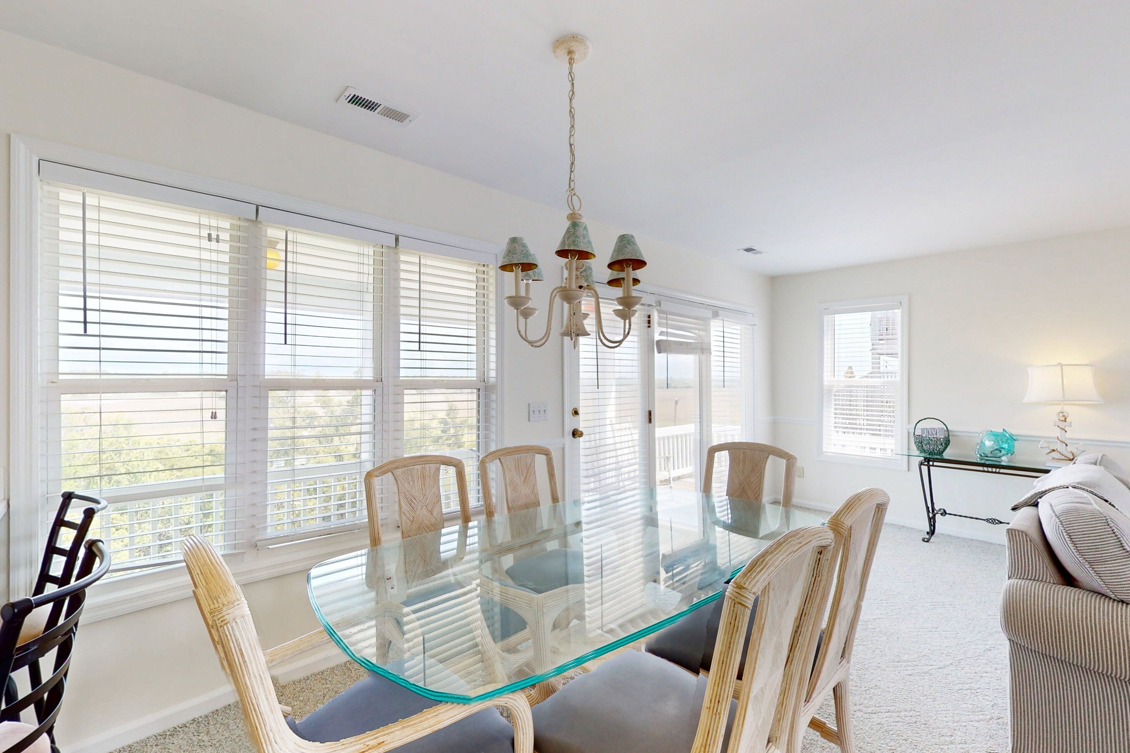 Un comedor con una mesa de vidrio rodeada de sillas, ventanas amplias con persianas y un candelabro.