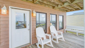 A wooden deck with a white door, two white chairs, and two lantern-style lights. - Shiloh by the Sea: Ocean-View Home with Easy Beach Access, WiFi & Central AC (Holden Beach)
