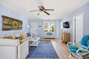 Un salón con ventilador de techo, un sofá, una mesa de centro, un tapete azul estampado, una silla de mimbre y un televisor montado en la pared.