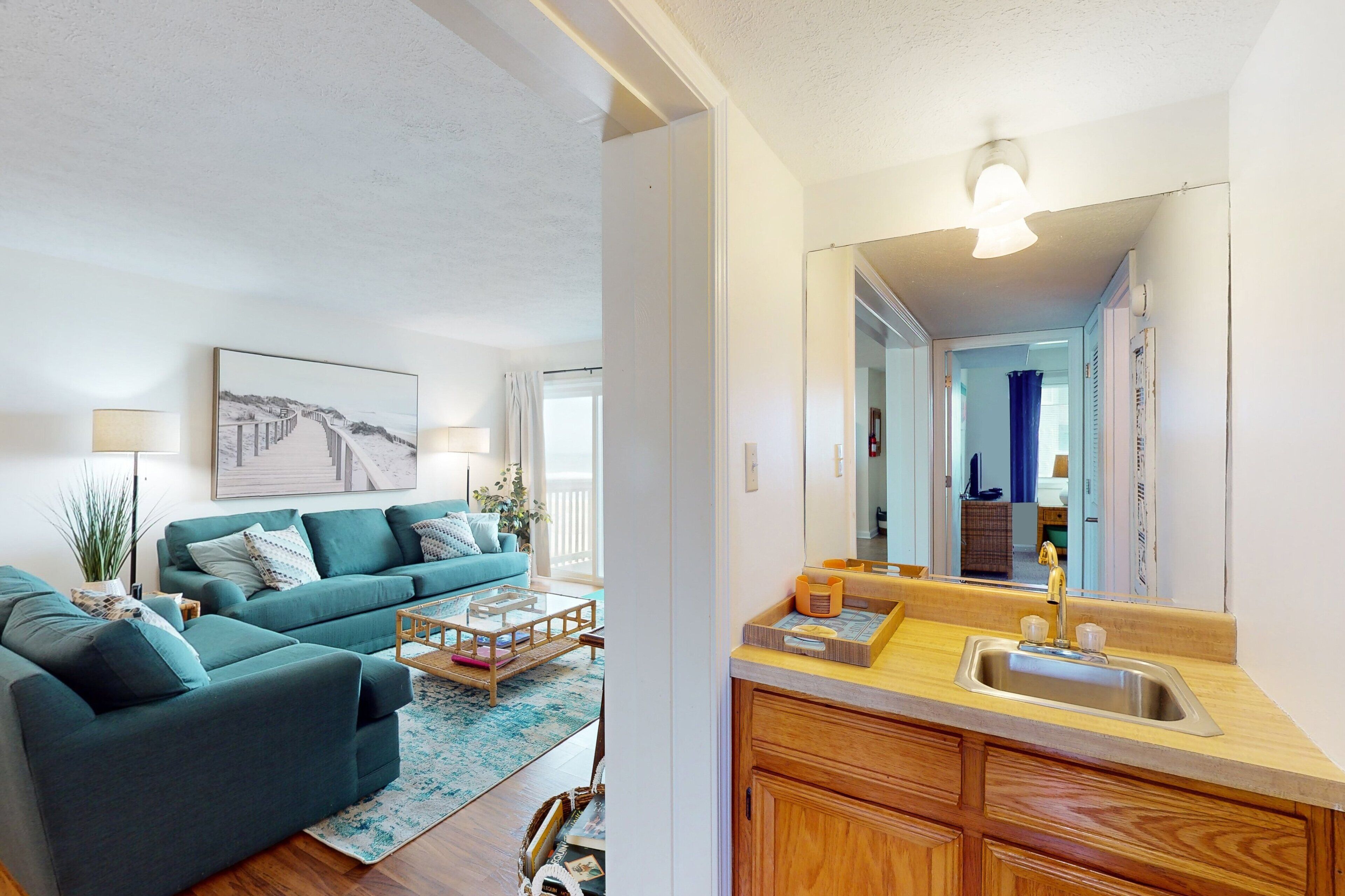 A modern living room with a teal sofa, a coffee table, and a large mirror above the kitchen sink.