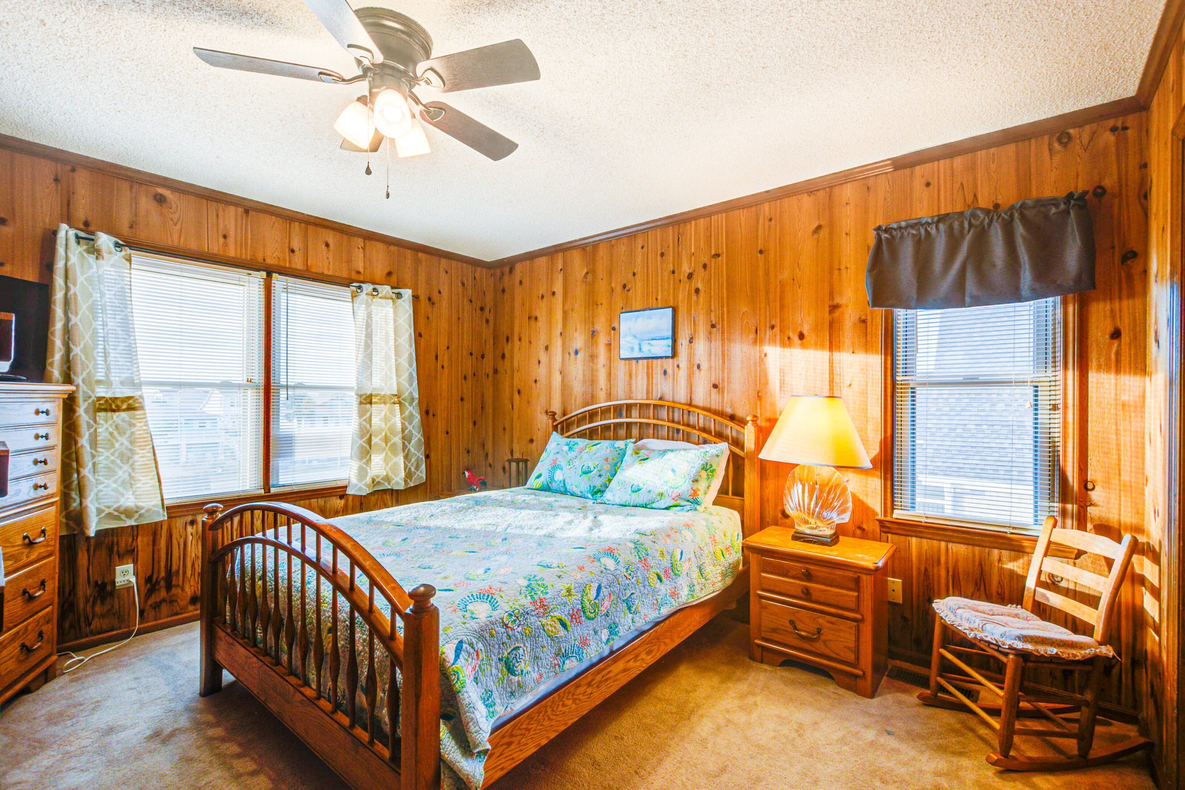 A wooden-paneled bedroom with a bed, bedside table, chair, and a ceiling fan.