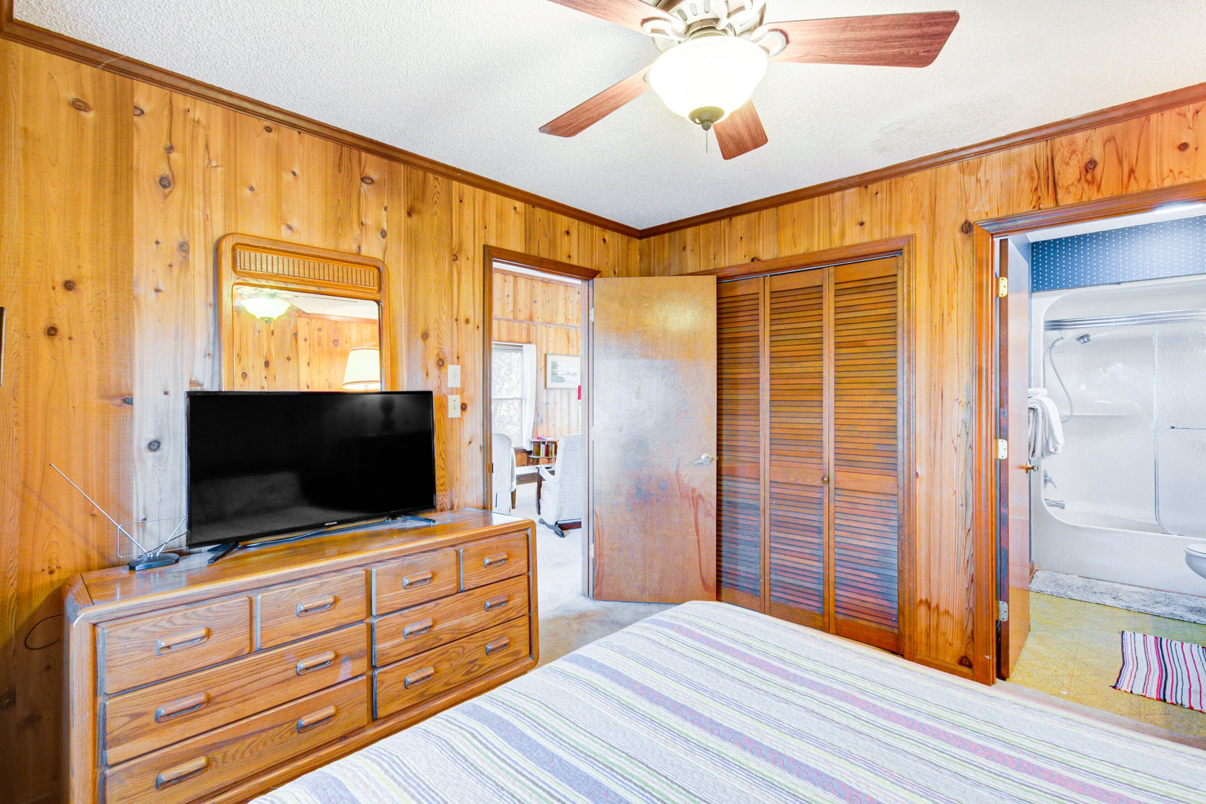 Un dormitorio con paneles de madera, un ventilador de techo, un televisor sobre una cómoda, una cama con colcha a rayas y una puerta corrediza que da a un baño.