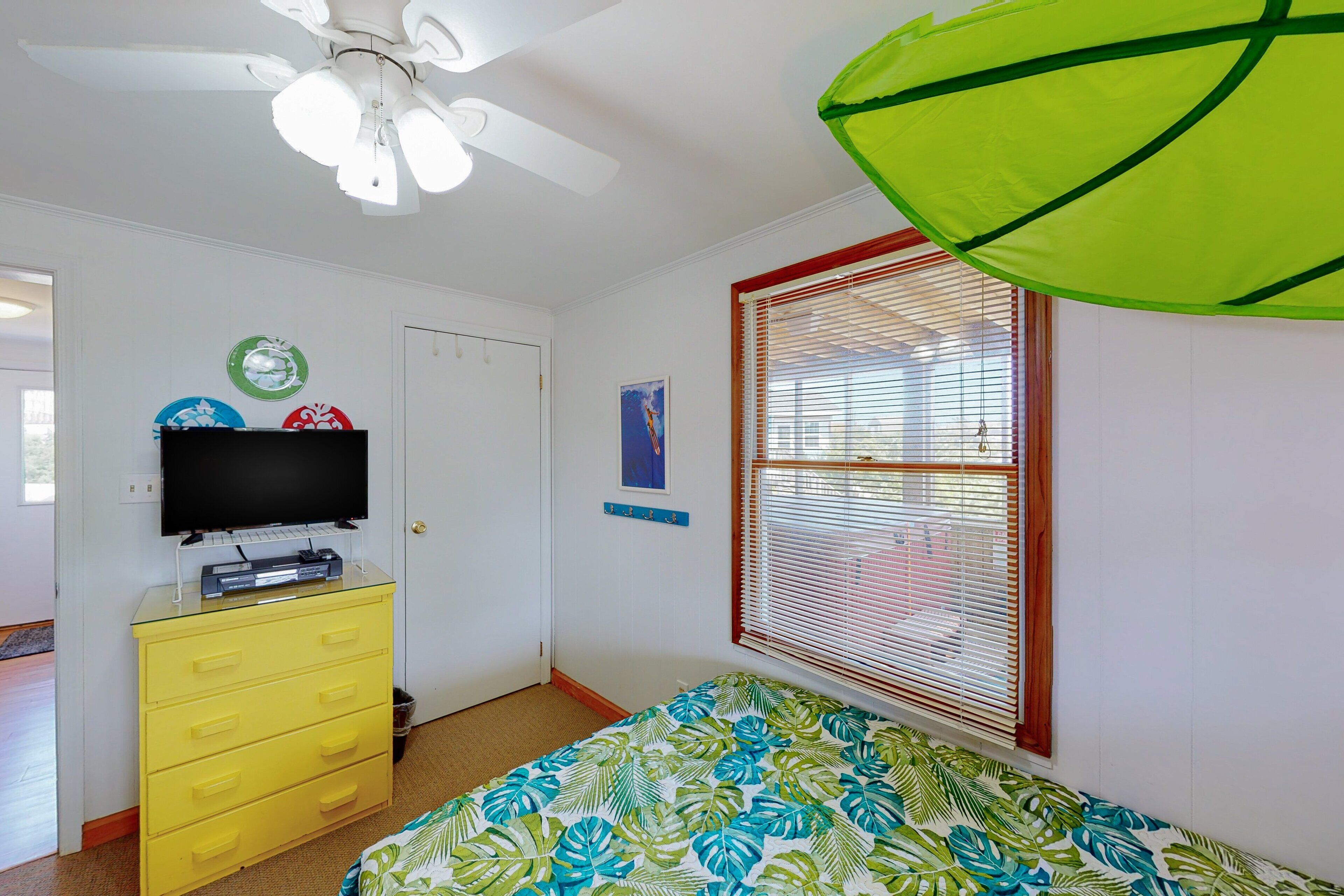 Une chambre à coucher avec un lit, un ventilateur de plafond, un téléviseur posé sur une commode et une fenêtre avec des stores.