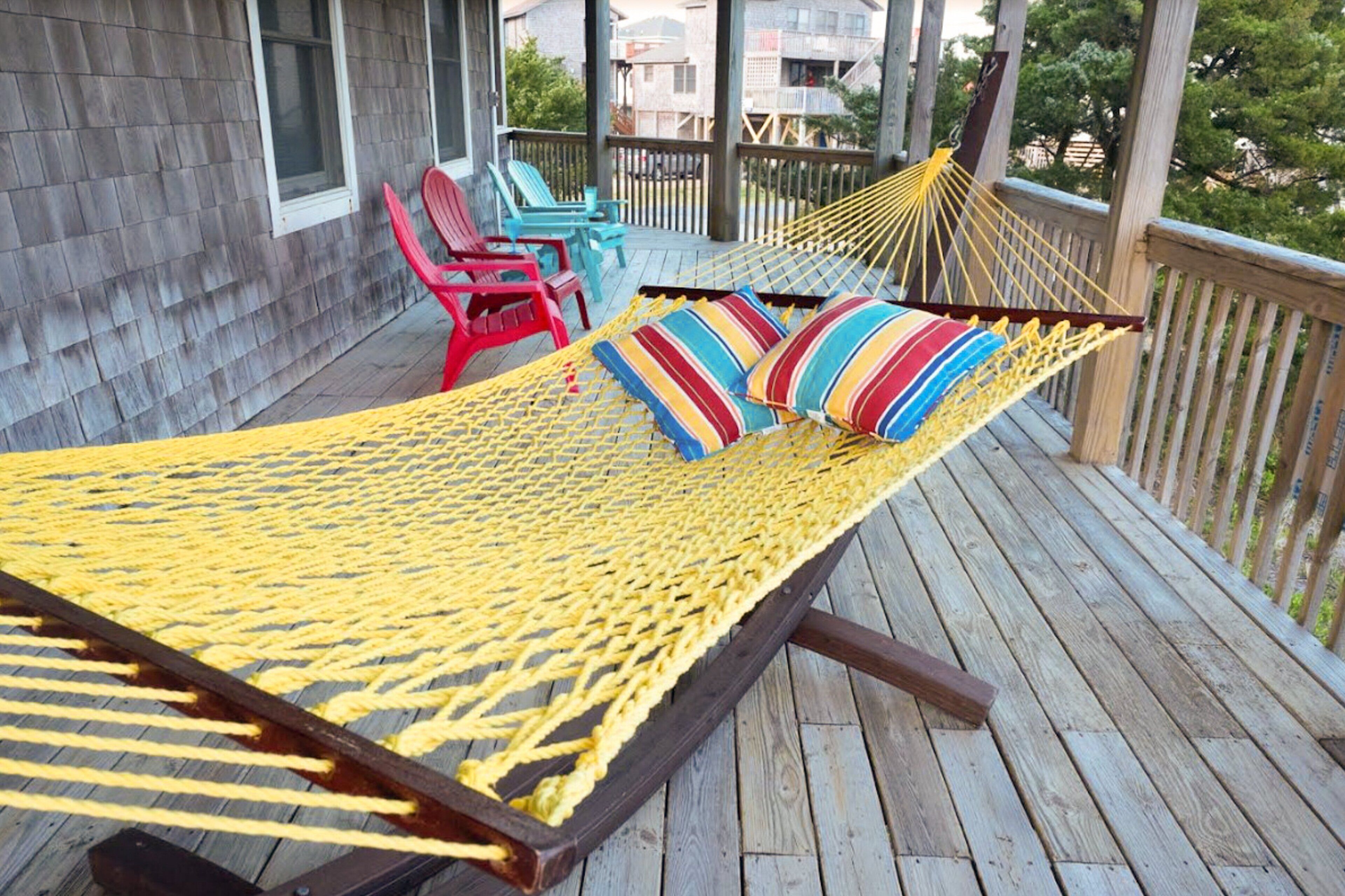 A hammock on a wooden deck with colorful pillows, red and blue chairs, and a wooden building in the background.