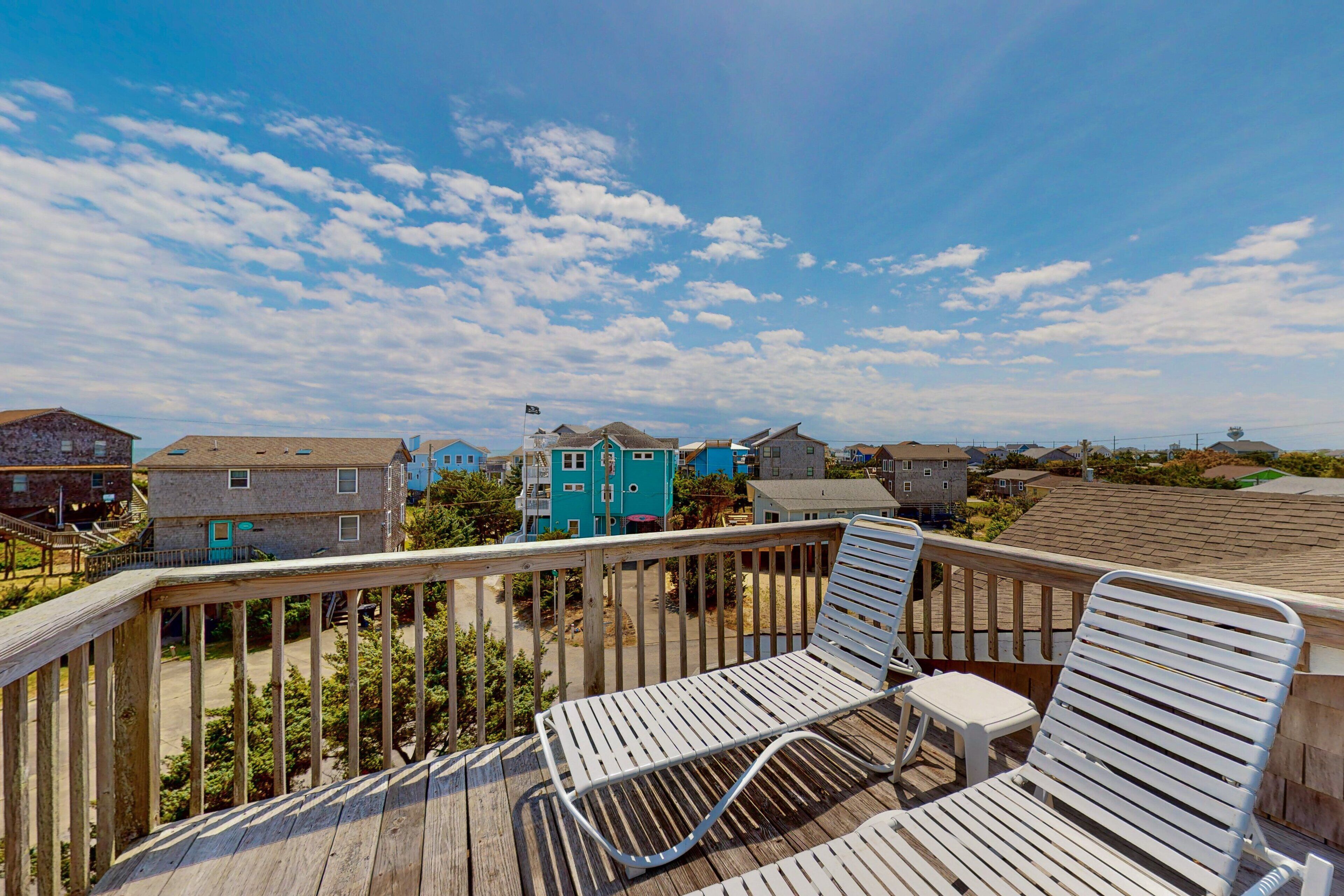 A wooden deck with two white lounge chairs, overlooking a residential area with colorful houses and a clear sky.