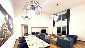 A modern dining area with a long table set for six, surrounded by dark blue chairs. There is a sofa set in the background and a large window with blinds.