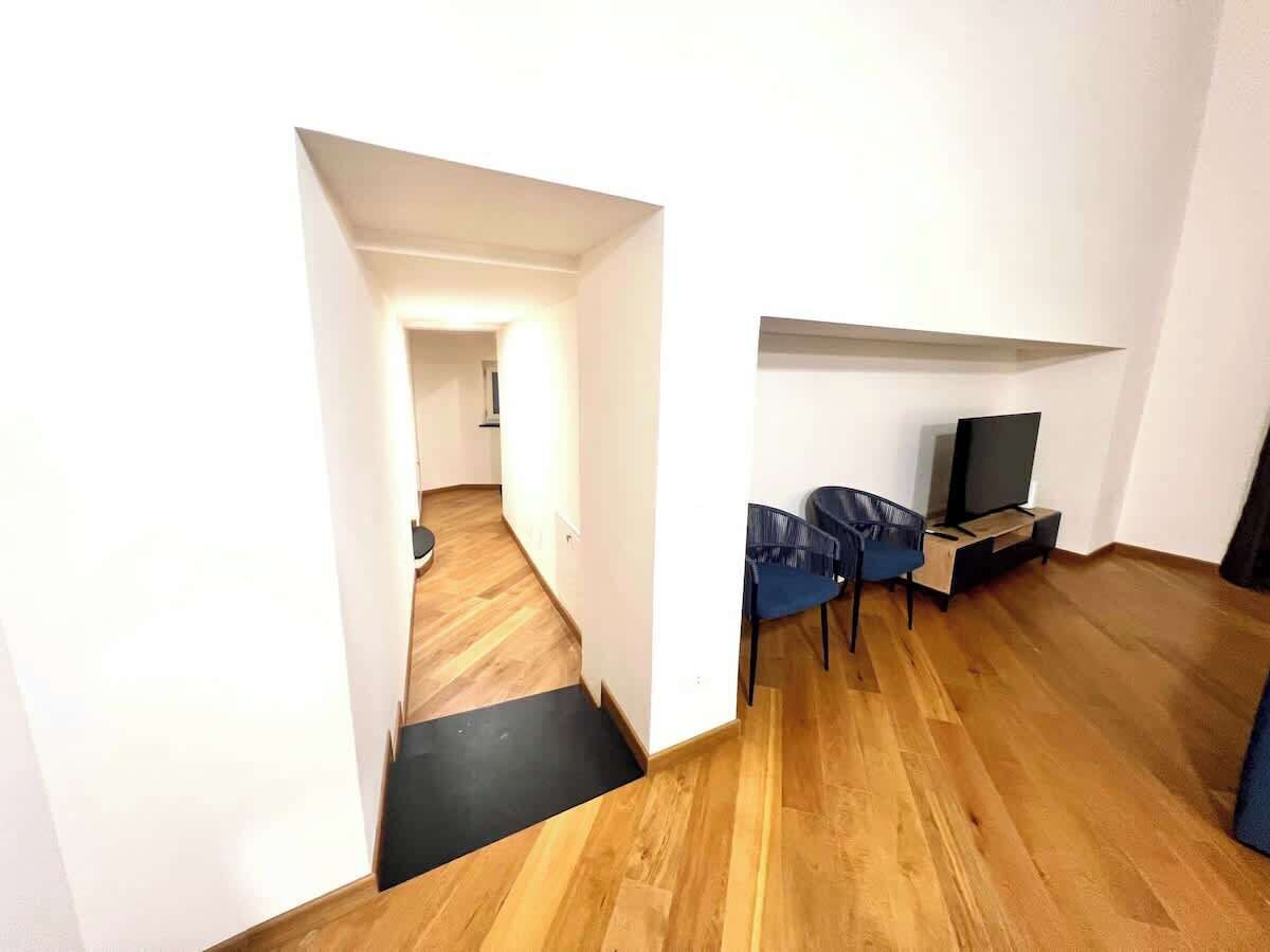 A modern living room with a wooden floor, a black mat at the entrance, a TV stand with a flat-screen TV, and two blue chairs.