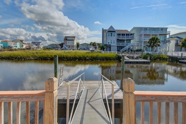 A waterfront property with a wooden dock leading to a calm water body, surrounded by residential buildings with balconies and a clear sky. - Canal-Front Home & Porch - Floating Boat Dock, Central AC, Outdoor Shower, WiFi (Holden Beach)