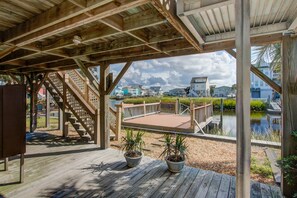 A wooden deck with potted plants, a staircase, and a view of a water body and buildings in the background. - Canal-Front Home & Porch - Floating Boat Dock, Central AC, Outdoor Shower, WiFi (Holden Beach)