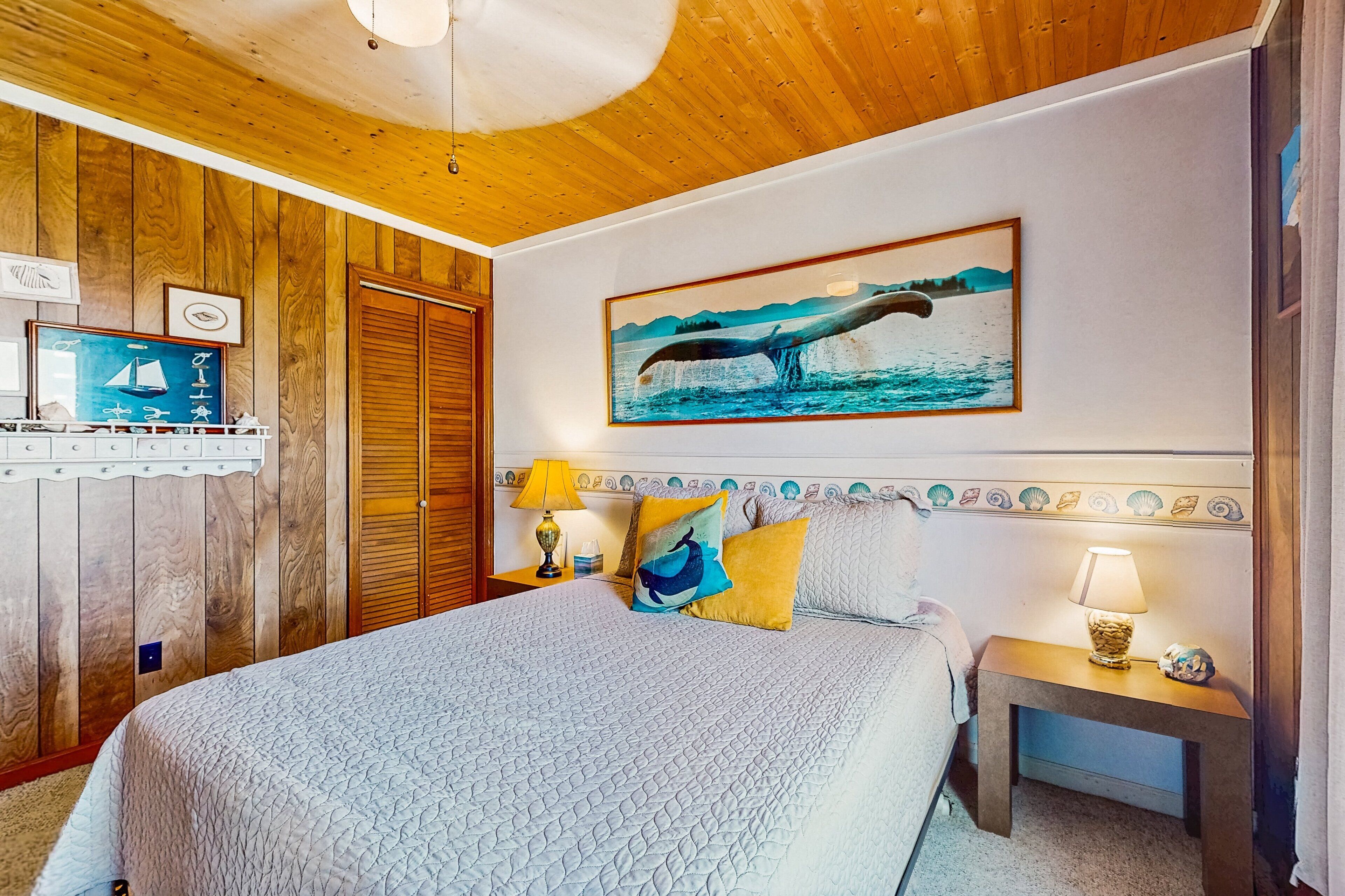 Une chambre avec un plafond en bois, un lit recouvert d’une courtepointe matelassée, un tableau représentant une baleine accroché au mur et une table de chevet avec une lampe.