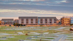 A residential complex with multiple apartment buildings, a central courtyard with picnic tables, and a clear sky. - Ocean view condo with pool access, furnished balcony, & central AC (Rodanthe)