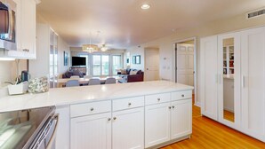 A modern kitchen with white cabinetry, a central island, and a dining area in the background. - Ocean view condo with pool access, furnished balcony, & central AC (Rodanthe)