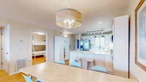 A modern kitchen with white cabinets and a central island, a dining area with a long table and blue chairs, and a bunk bed in the background. - Ocean view condo with pool access, furnished balcony, & central AC (Rodanthe)