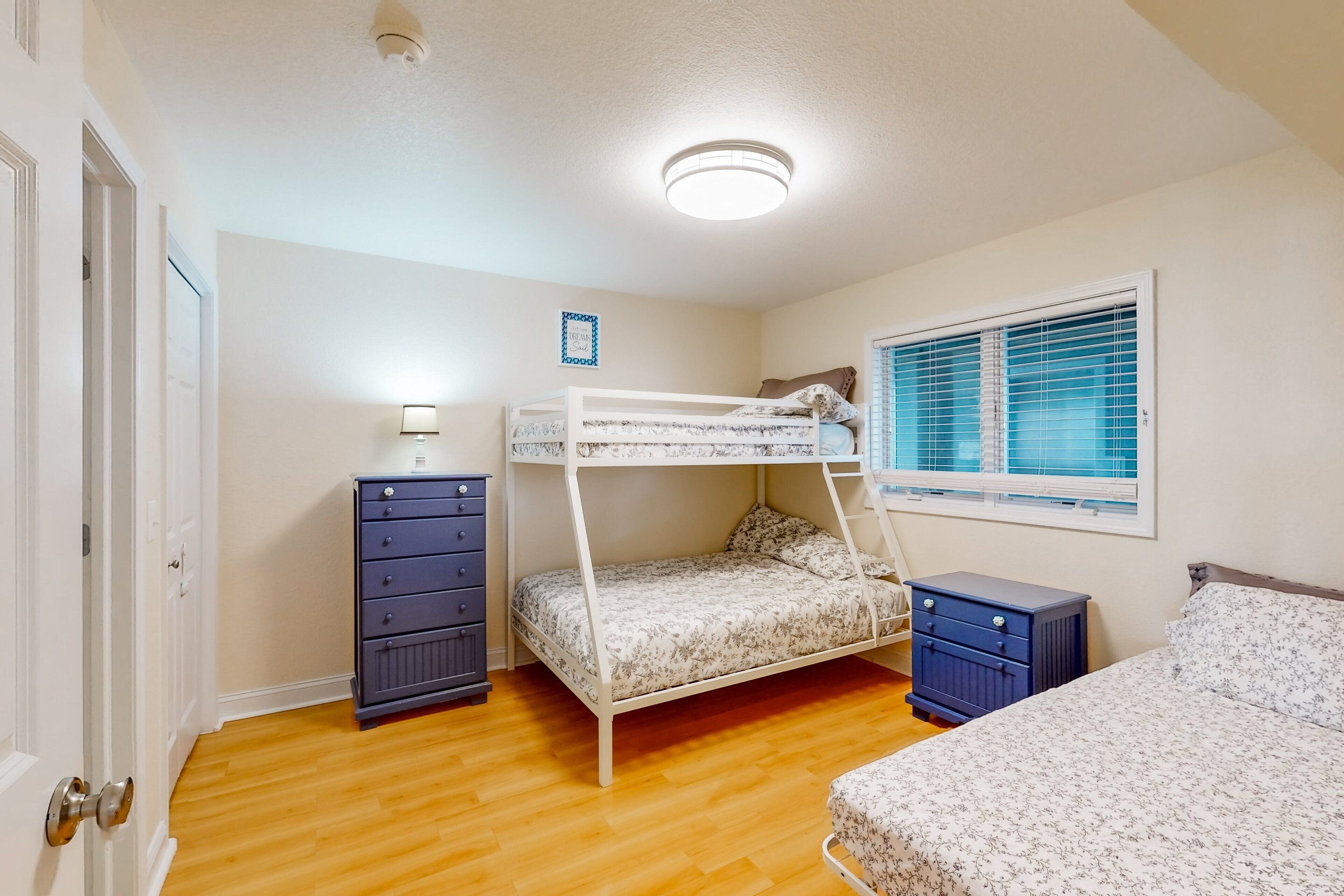 A bedroom with a bunk bed, a dresser, a bedside table, and a window with blinds.
