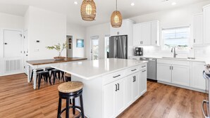 Una cocina moderna con armarios blancos, una isla grande y un refrigerador de acero inoxidable. Hay dos lámparas colgantes y una planta en maceta sobre la isla.