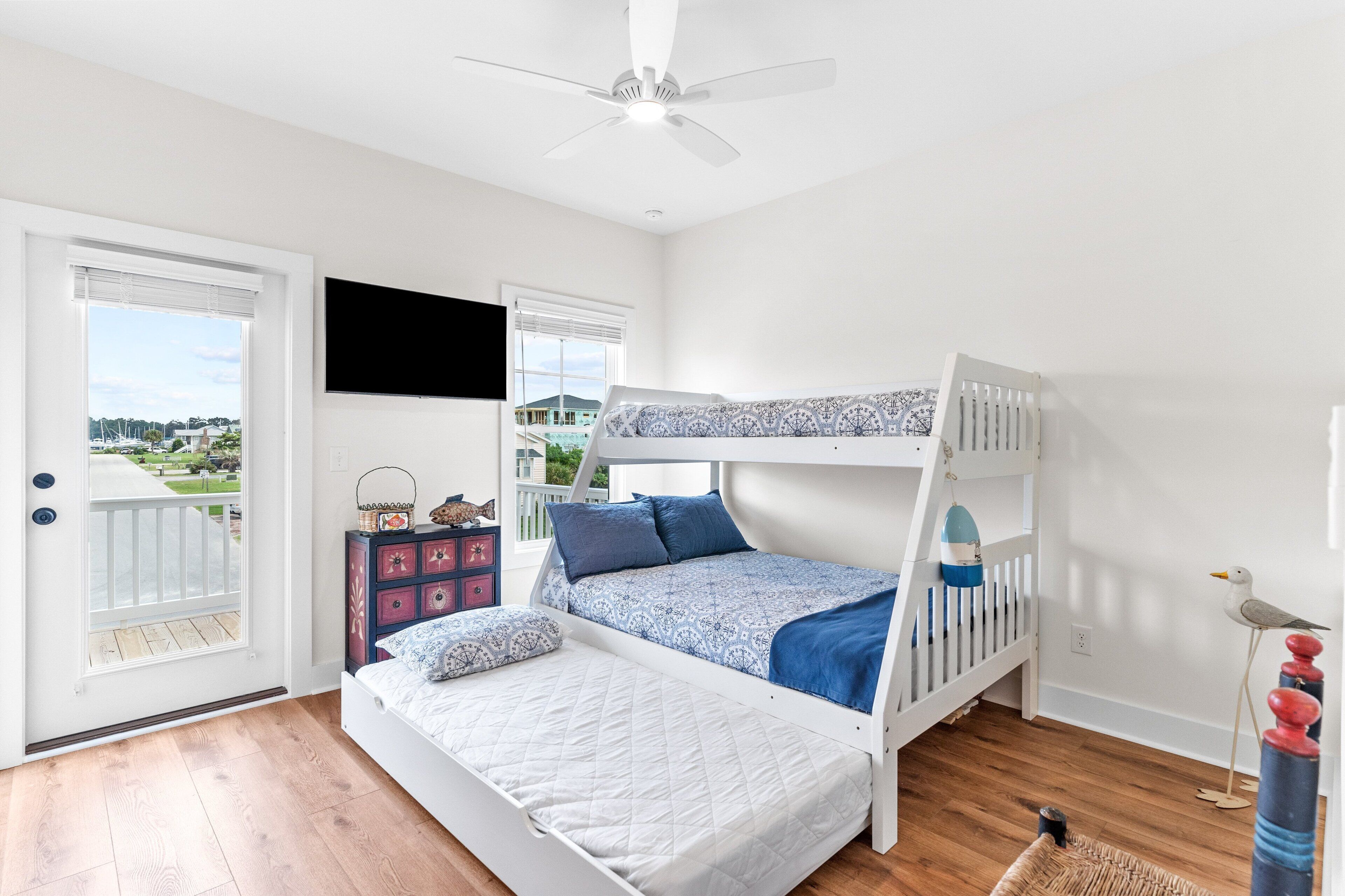 A bedroom with a bunk bed, a desk, and a chair. There is a window with a view of a house and a balcony.