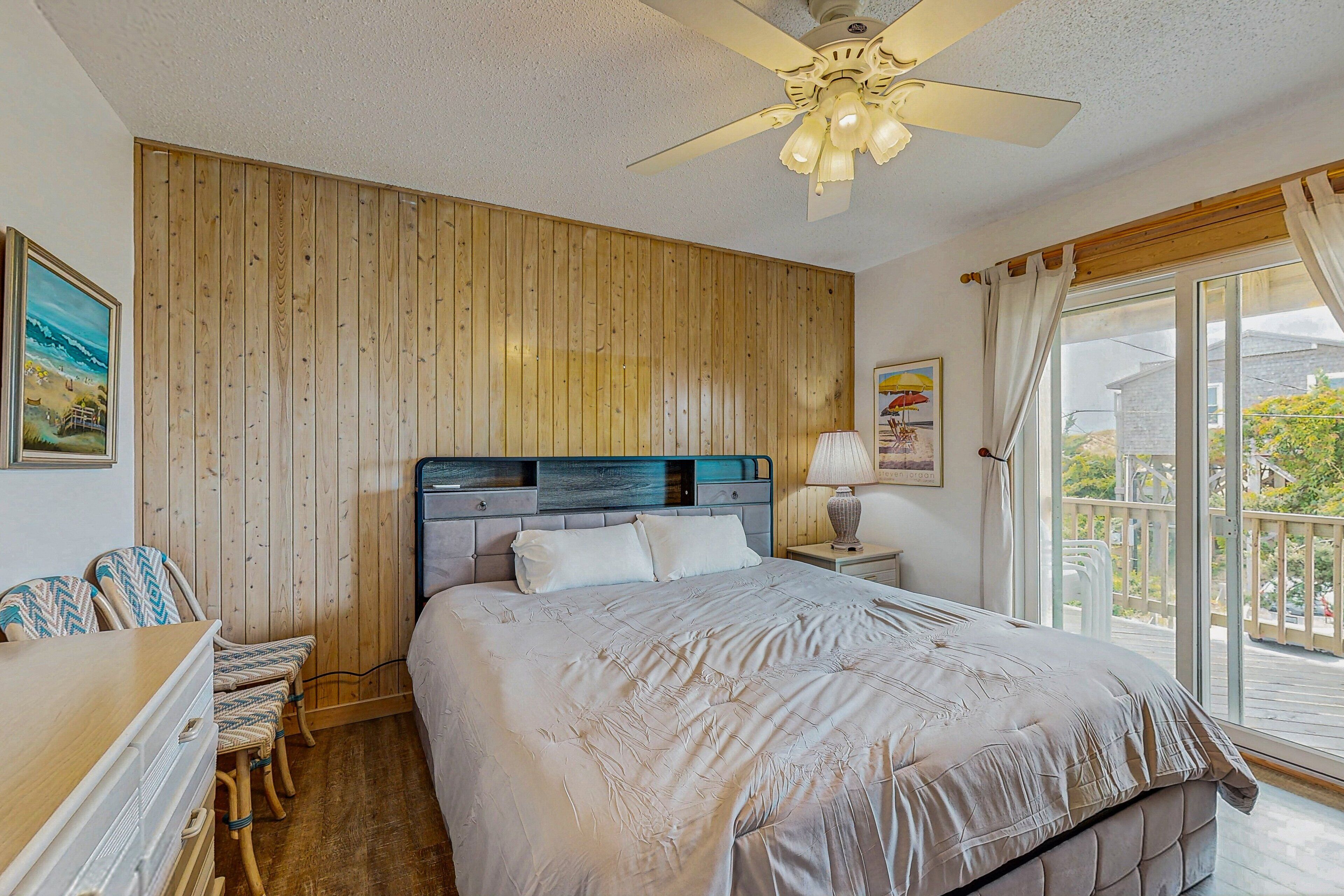 A bedroom with a wooden headboard, a bed with a white bedspread, a desk with chairs, and a balcony with a view of the outside.