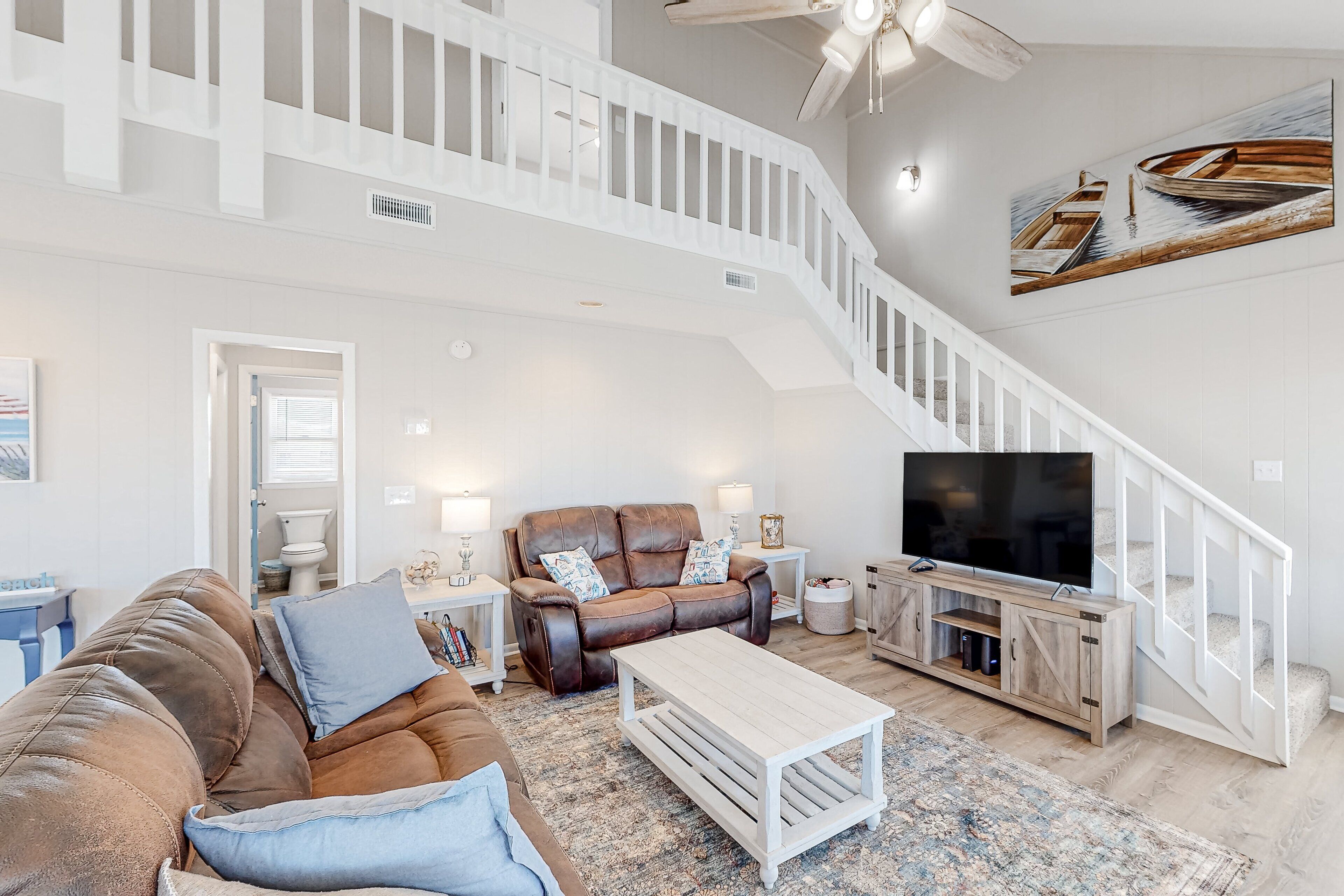 A spacious living room with a brown leather sofa, a white coffee table, and a TV on a wooden stand. A staircase with a white railing leads to an upper level.