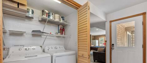 Un área de lavandería con dos lavadoras blancas, estantes con diversos objetos y una puerta de madera.