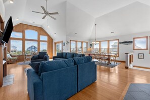 A spacious living room with a large blue sectional sofa, a dining area with a table and chairs, and large windows offering views of the outside.