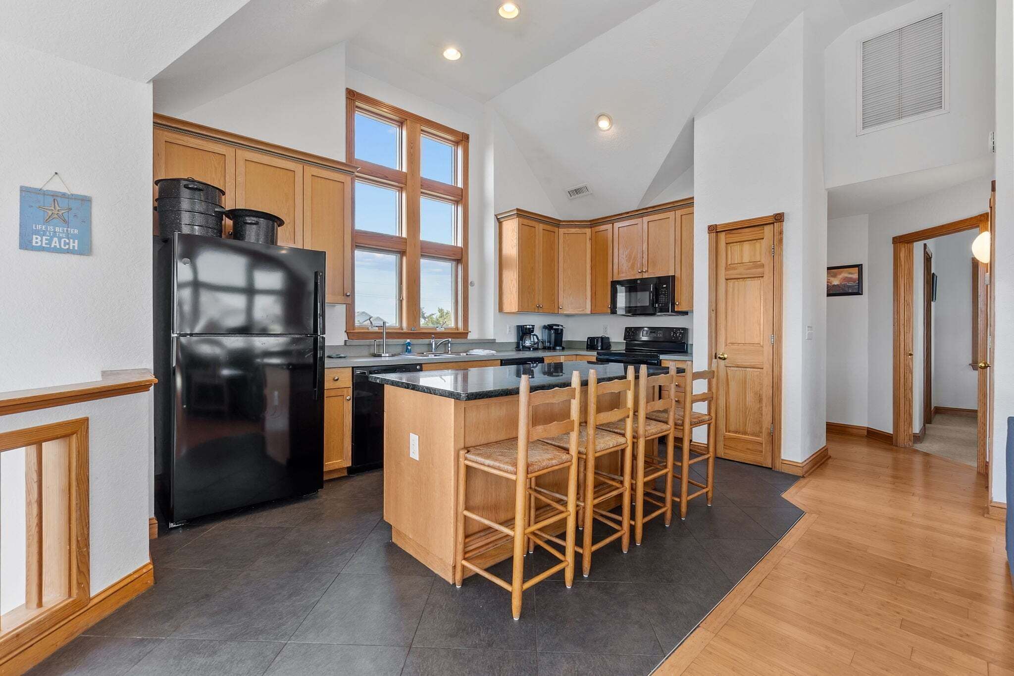 A modern kitchen with wooden cabinets, a black refrigerator, a coffee station, and a large window.
