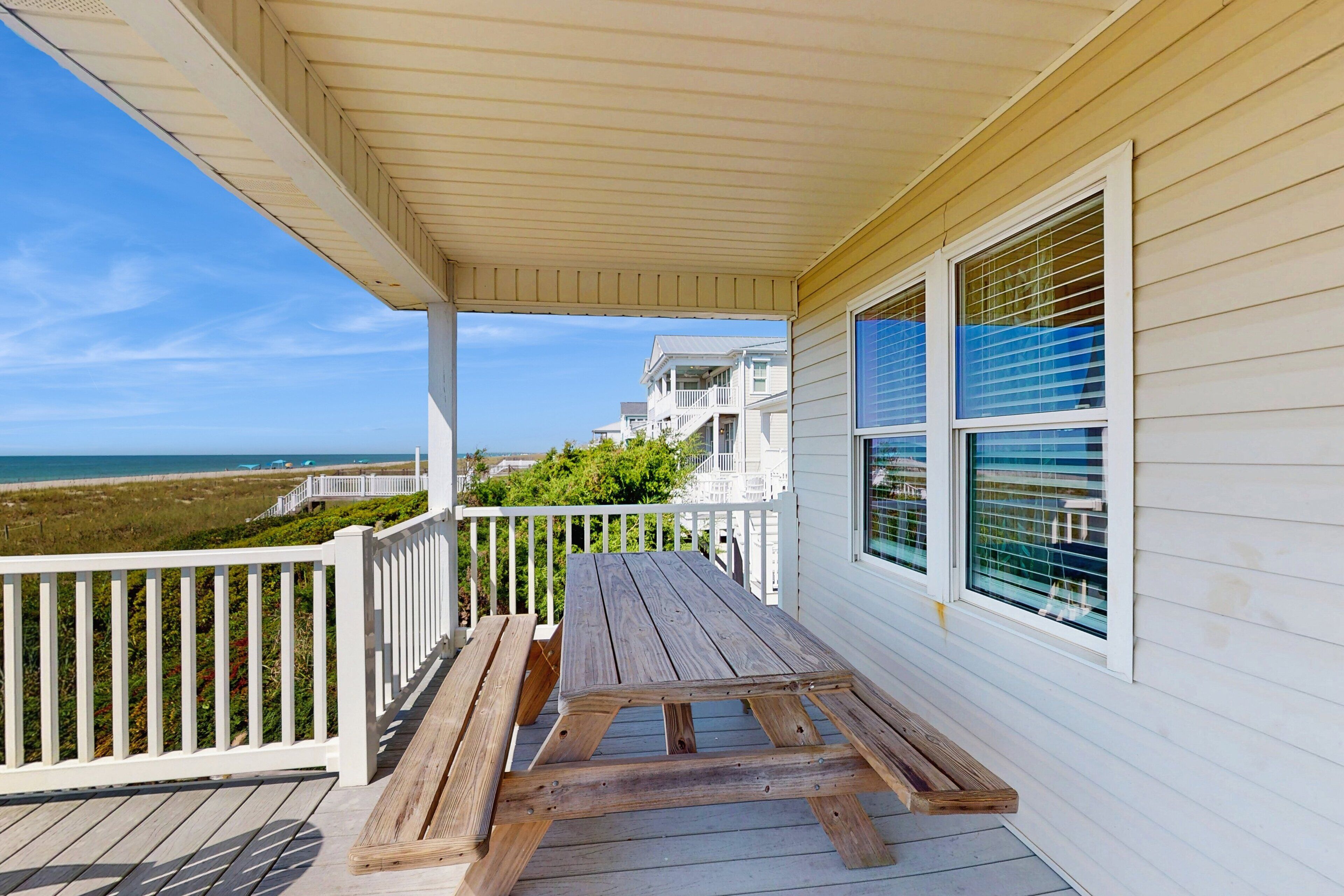 A wooden deck with a picnic table, white railings, and a view of the ocean and beach.
