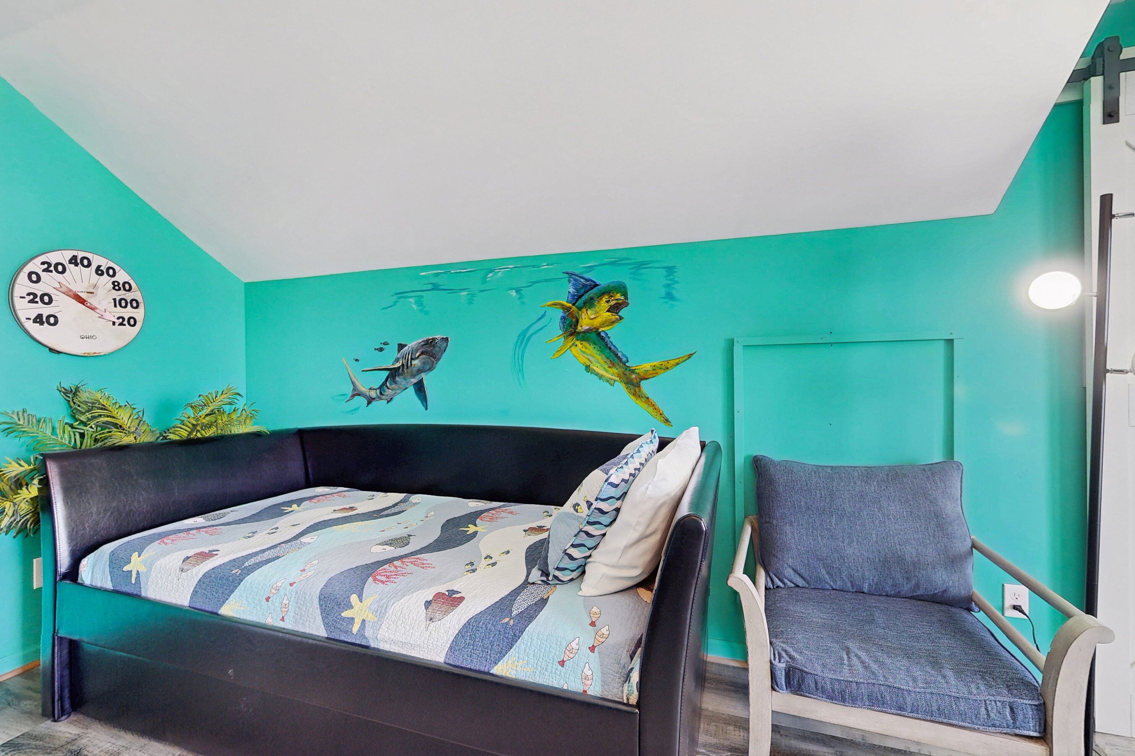A room with a teal wall, a bed with a colorful quilt, a chair, and a wall clock.