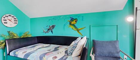 A room with a teal wall, a bed with a colorful quilt, a chair, and a wall clock.