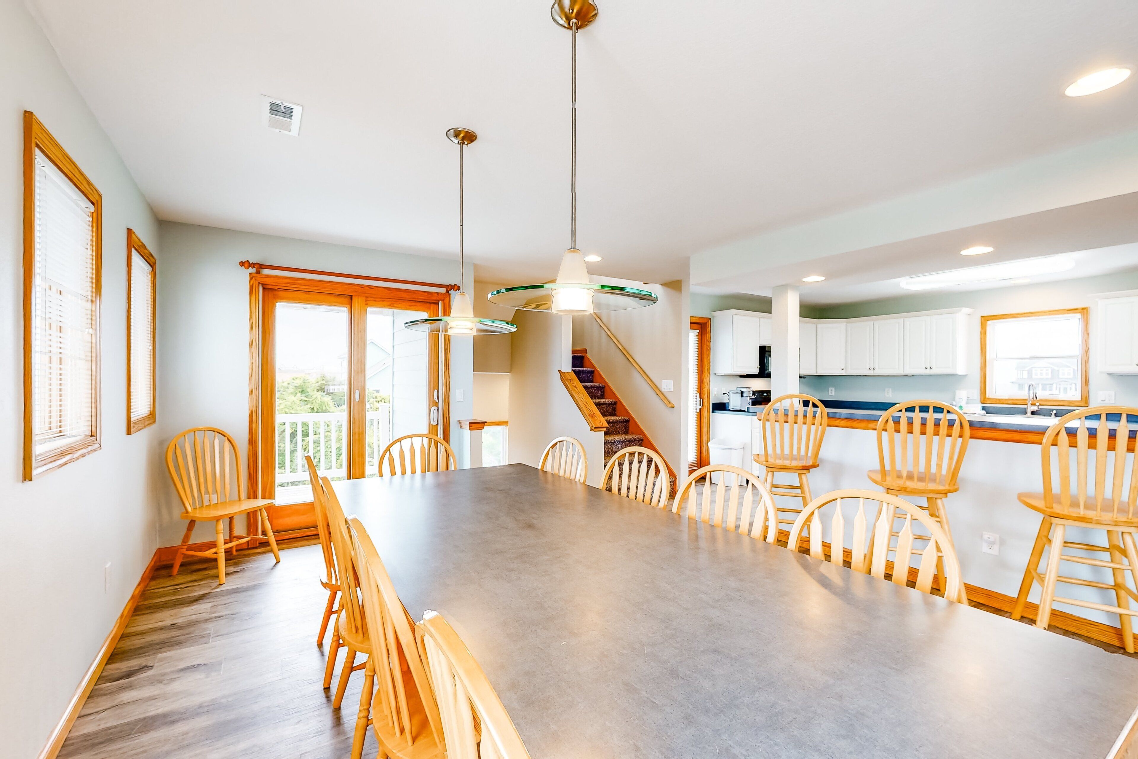A spacious dining area with a large table and wooden chairs, a kitchen with white cabinets, and a staircase leading to an upper level.