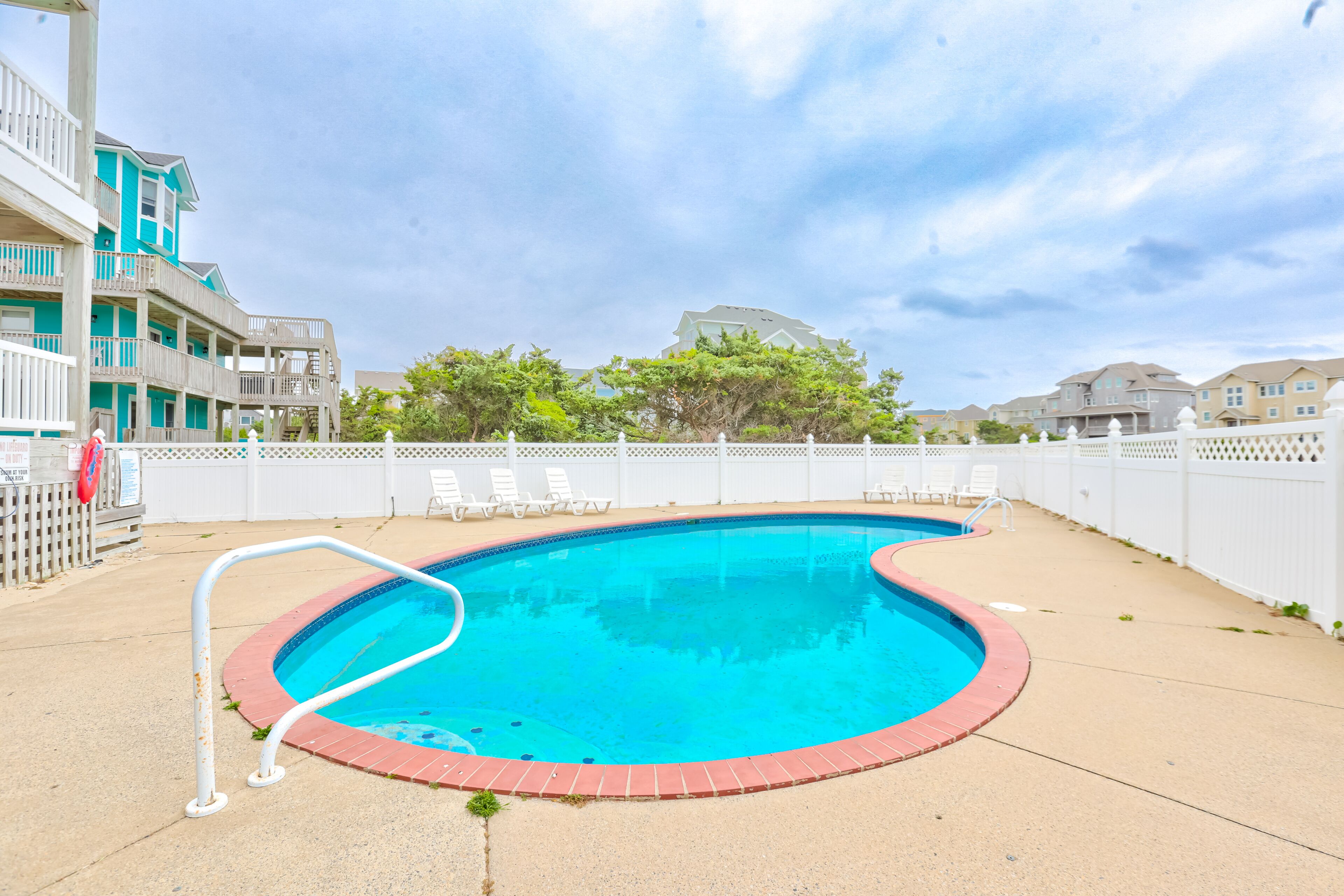 A swimming pool with a white deck, surrounded by a white fence, and adjacent to a building with multiple balconies.