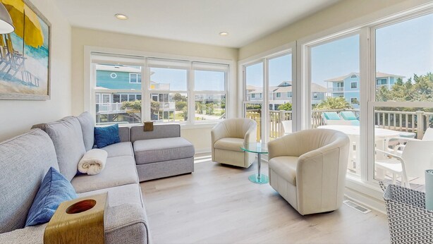 A spacious living room with a grey sofa, a glass coffee table, and a beige armchair. Large windows offer a view of neighboring houses and a balcony.