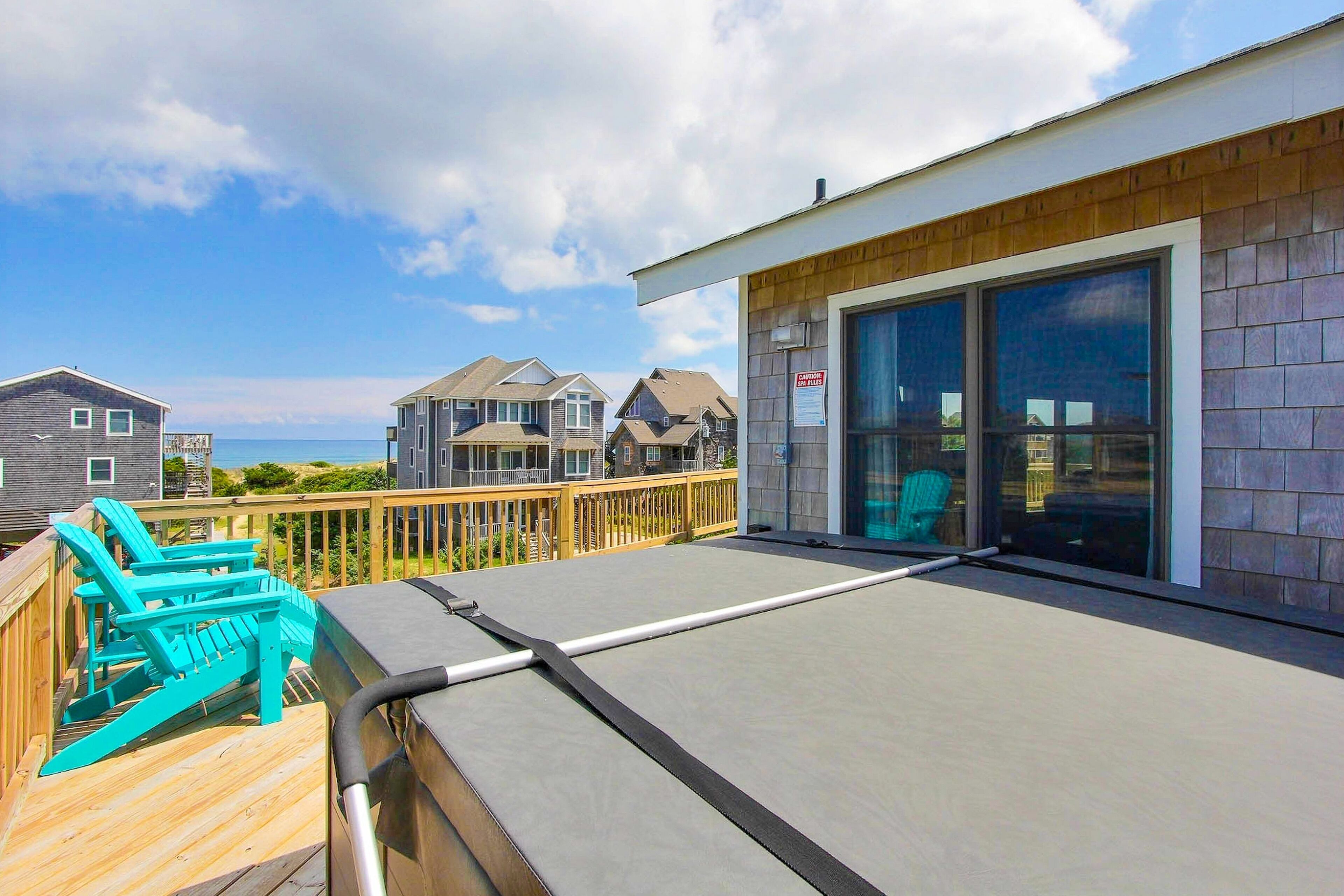 A deck with a grill and Adirondack chairs, overlooking a residential area with houses and a clear sky.