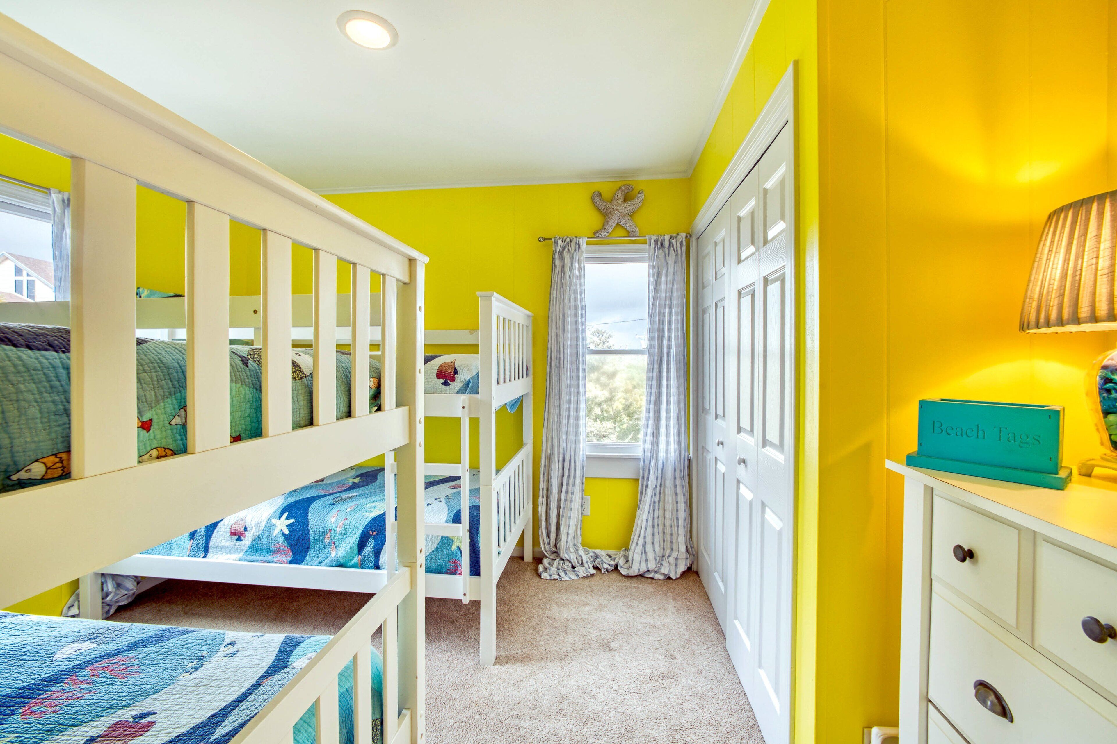 A child's bedroom with bunk beds, a window with curtains, a dresser, and a lamp.