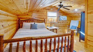 Un dormitorio de cabaña de madera con una cama, ventilador de techo, ventana, silla y refrigerador.