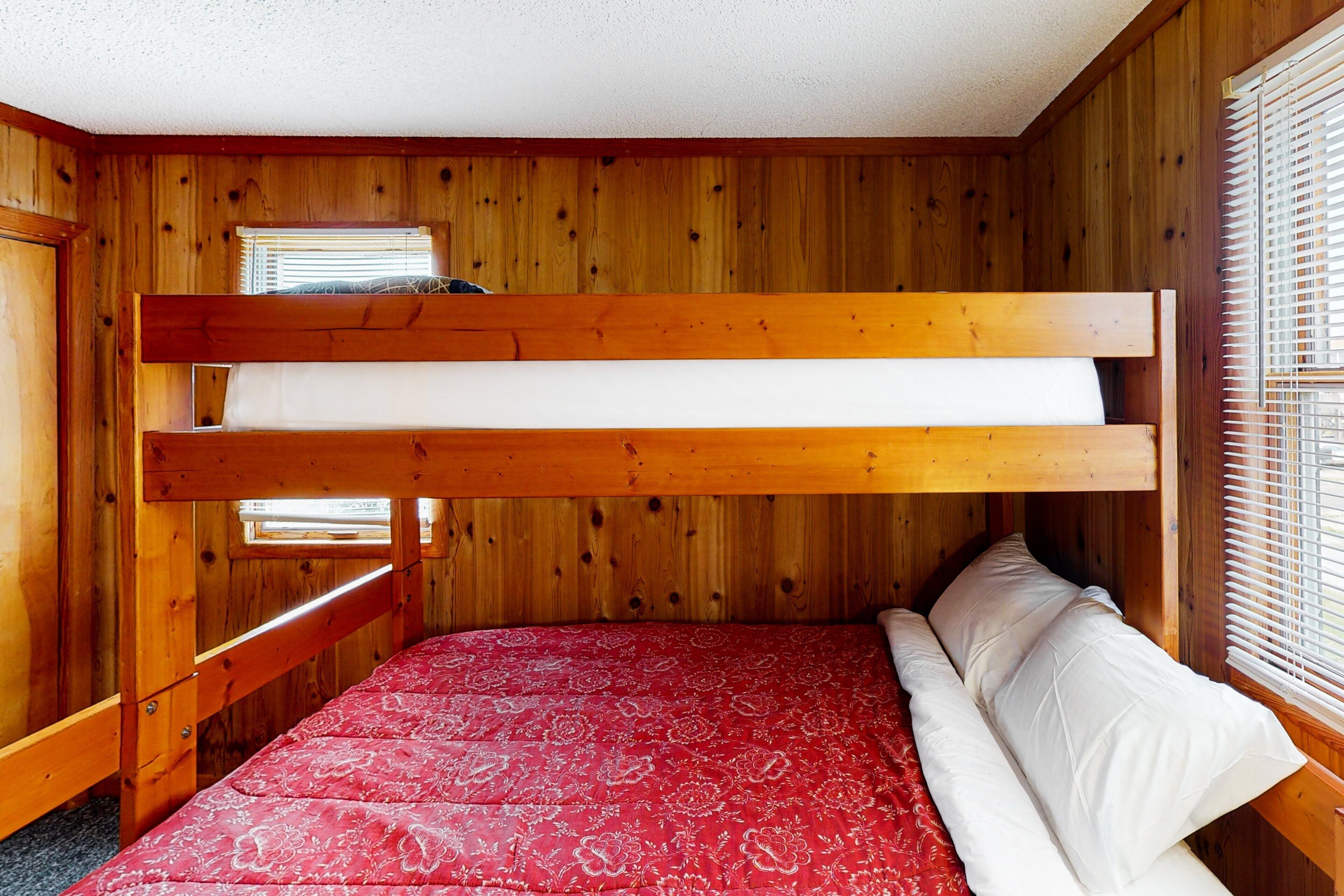 Una cama litera con un edredón rojo estampado y almohadas blancas en una habitación de madera con ventana y persianas.