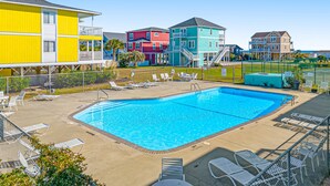 A swimming pool with lounge chairs and a fenced area, surrounded by colorful buildings. - Condo near the beach with pool, BBQ area, balcony, & fireplace (Holden Beach)