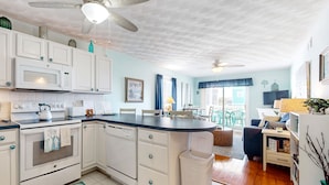 A kitchen with white cabinets, a microwave, and a dishwasher. There is a dining area with a table and chairs, and a ceiling fan. - Condo near the beach with pool, BBQ area, balcony, & fireplace (Holden Beach)