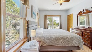 Un dormitorio con cama, mesita de noche, lámpara y vistas a los árboles por la ventana.