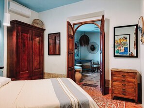 Un dormitorio con cama, armario de madera y cómoda. Hay un espejo en la pared y una puerta que da a otra habitación.