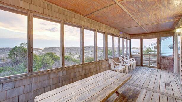 Une terrasse en bois avec un banc et des chaises, de grandes fenêtres offrant une vue sur le paysage.