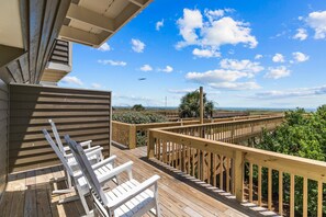 Una terraza de madera con sillas blancas y barandilla, con vista a una playa con palmeras y cielo despejado.