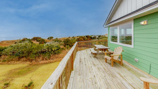 Une terrasse en bois avec une table et des chaises, donnant sur une pelouse agrémentée d’arbustes et offrant une vue lointaine sur l’océan.