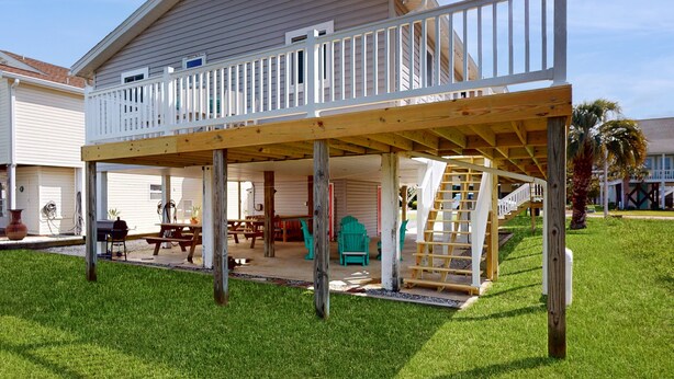 A two-story house with a covered patio area, a wooden deck, and a staircase leading to the upper floor. - Bright Home with Deck, Patio, High-Speed WiFi, W/D, & Central AC - Near Beach (Holden Beach)