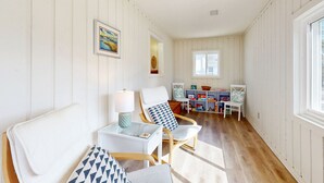 A room with white paneled walls, a wooden floor, a white armchair with a blue pillow, a small table with a lamp, and a bookshelf in the background. - Bright Home with Deck, Patio, High-Speed WiFi, W/D, & Central AC - Near Beach (Holden Beach)