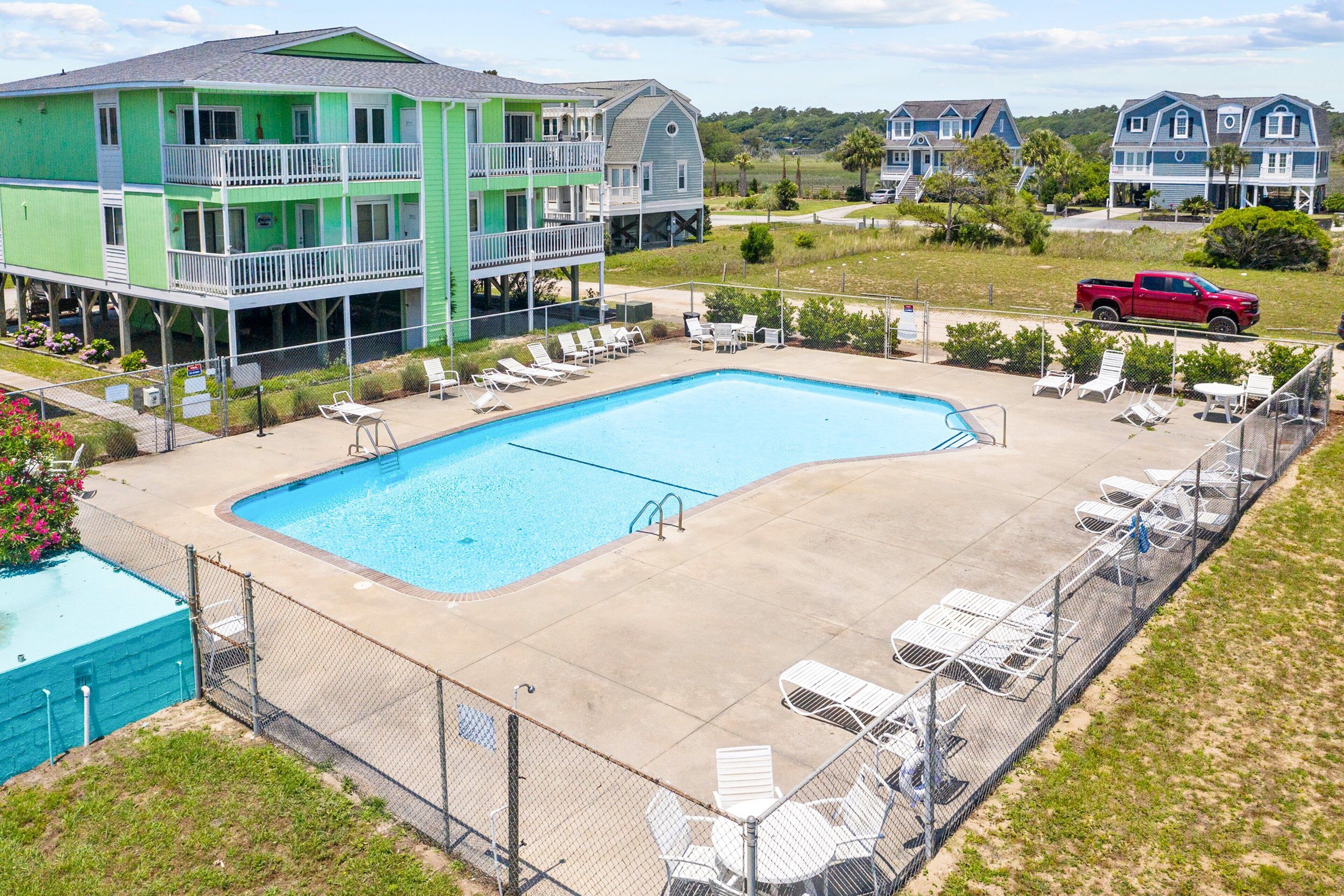 Une piscine avec des chaises longues, une terrasse entourée d’une clôture et un immeuble à plusieurs étages doté de balcons.